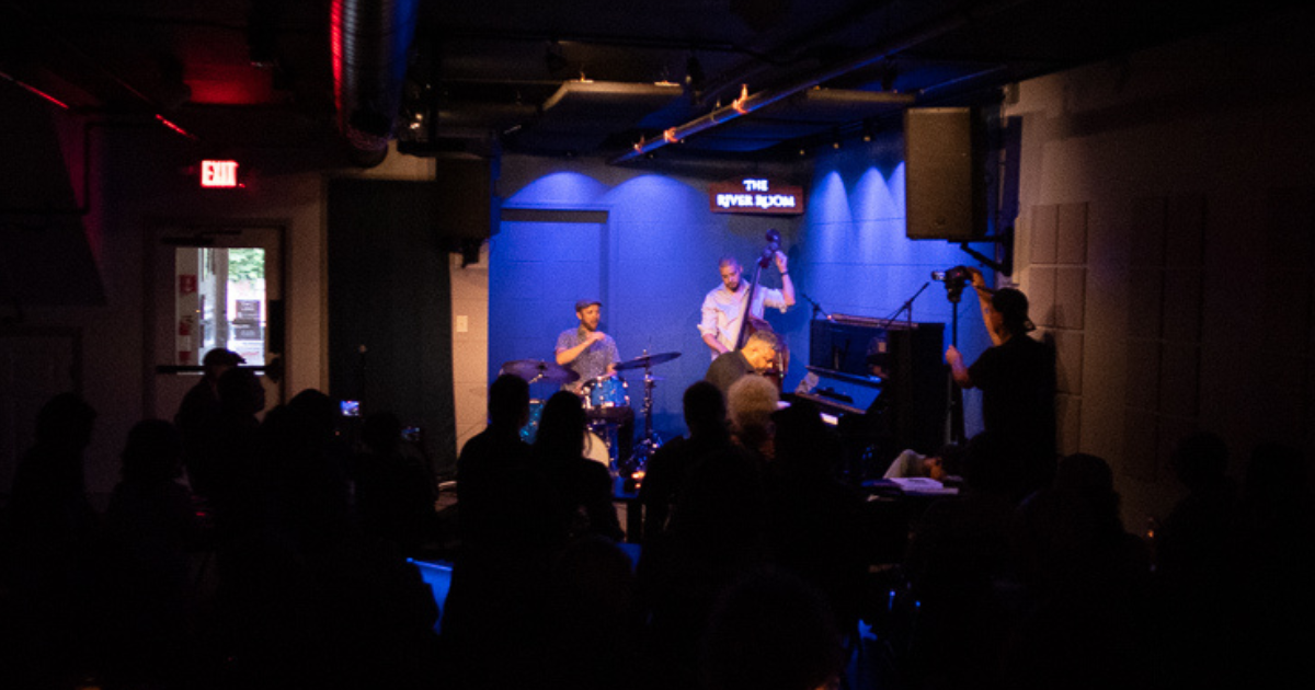 Zaccai Curtis on piano, Luques Curtis on upright bass, and Richie Barshaw on drums perform on a blue-lit stage at The River Room.