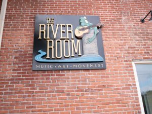 The River Room sign on a brick wall, featuring a frog playing guitar and the words “Music · Art · Movement.”