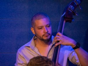 Luques Curtis plays upright bass at The River Room, his left hand positioned on the fingerboard under blue stage lighting.