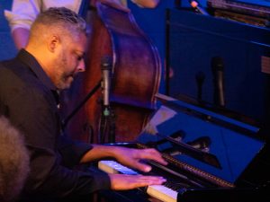 Zaccai Curtis plays an upright piano at The River Room, with his hands on the keys and an upright bass behind him.